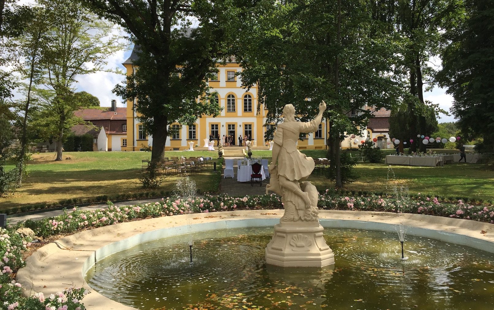 Der Schlossparkbrunnen befindet sich im Vordergrund. Eine Statue ragt aus dem Brunnen heraus und hat den Blick auf das Schloss gerichtet. Das Schloss Jaegersburg steht im Hintergrund, die Terasse ist festlich geschmückt.