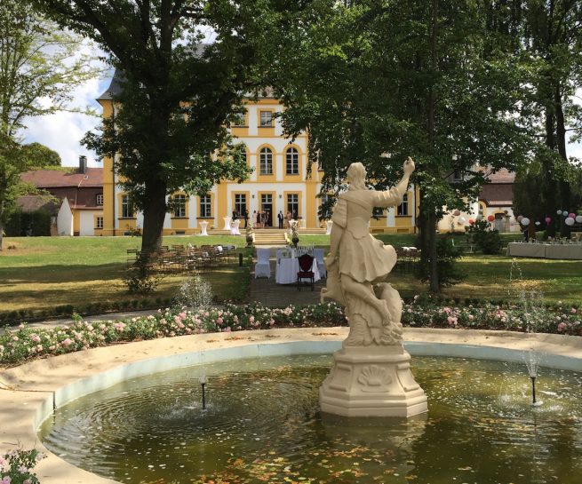 Der Schlossparkbrunnen befindet sich im Vordergrund. Eine Statue ragt aus dem Brunnen heraus und hat den Blick auf das Schloss gerichtet. Das Schloss Jaegersburg steht im Hintergrund, die Terasse ist festlich geschmückt.