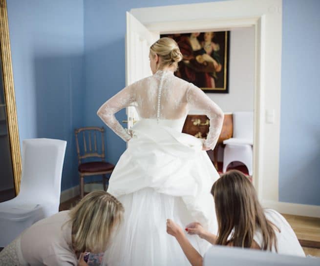 Die Trauzeuginnnen drappieren das Hochzeitskleid der Braut richtig. Die Braut ist mit dem Rücken zugewandt, während sie sich seitlich im Spiegel des Brautzimmers betrachtet. Das Hochtzeitskleid wird richtig ausgestellt