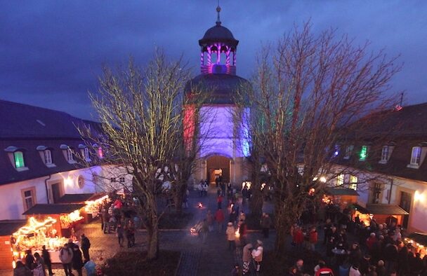 Der Weihnachtsmarkt im Schloss Jägersburg als Bild in Vogelperspektive. Menschen besuchen die im Hof aufgebauten leuchtenden Hütten.